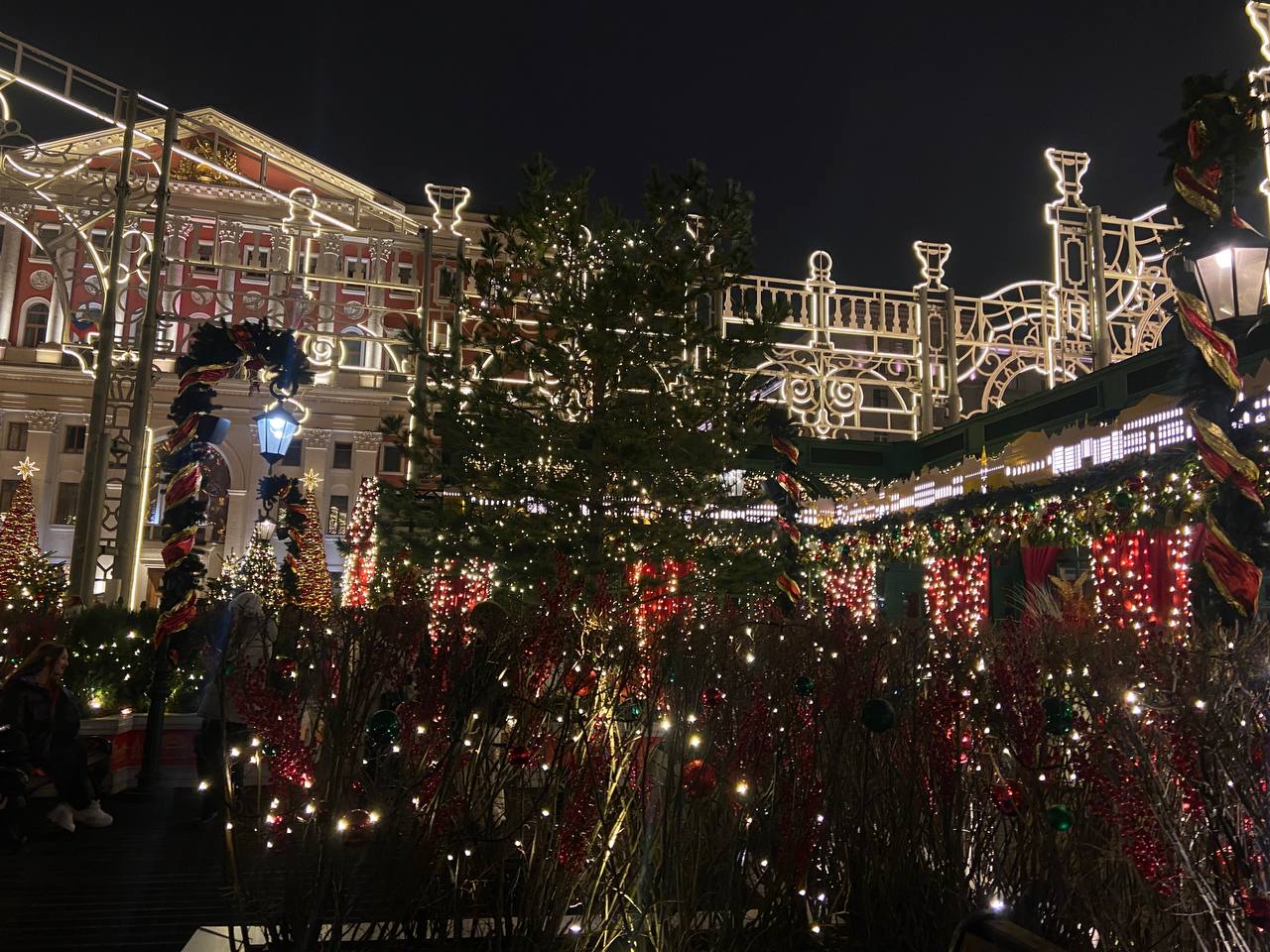 Яркие огни вечернего города освещают ели и красные декорации. Фото: Анастасия Петрова, «Вечерняя Москва»